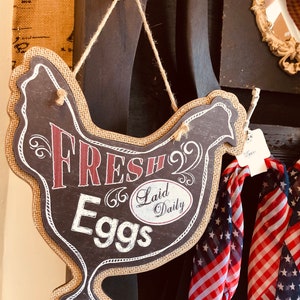 May include: A brown burlap chicken-shaped sign with the text "Fresh Laid Daily Eggs" in white and red lettering. The sign is hanging from a white knob on a wooden surface.