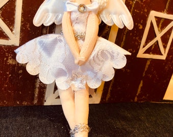 May include: A white fabric angel doll with a white lace dress, white wings, and a white bow around its neck. The doll has a white face with painted eyes and a rhinestone in the center of the bow.