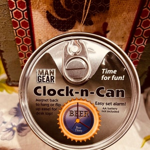 May include: A silver can clock with a black and white clock face and the words "Clock-n-Can" and "Time for fun!" printed on it. The can has a magnet on the back to hang it up and an alarm function. The clock face has a beer bottle cap design with the words "Beer Thirty" printed on it.