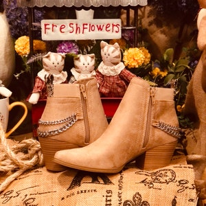 May include: A pair of tan suede ankle boots with a chunky heel and silver chain detail. The boots have a zipper on the inside of each ankle.
