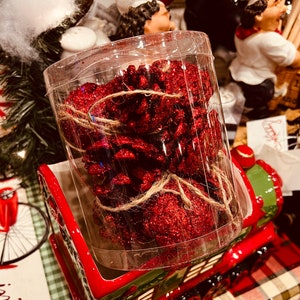 May include: A clear plastic container filled with red glitter pine cones tied with twine. The container is sitting on a red and green ceramic train.
