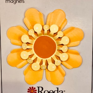 May include: A yellow flower-shaped magnet with a smaller orange flower in the centre. The magnet has the text "Roeda brighten your life" on the bottom.