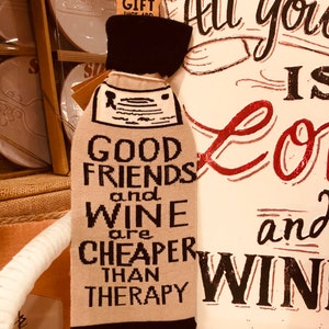 Könnte beinhalten: Beigefarbene Weinflaschenhülle mit schwarzem Besatz und dem Text "Good Friends and Wine are Cheaper Than Therapy".