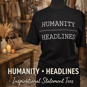 Puede incluir: Camiseta negra con las palabras "HUMANITY HEADLINES" en blanco sobre un maniquí. Forma parte de la colección "Inspirational Statement Tees". El fondo incluye una mesa de madera, velas y otras prendas.
