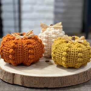 Op de afbeelding: Drie handgemaakte gehaakte pompoenen in oranje, crème en goud, geplaatst op een houten schijf. Elke pompoen heeft een textuur met bobbelsteek en is afgewerkt met een takje en raffia. Een rustieke herfstdecoratie.