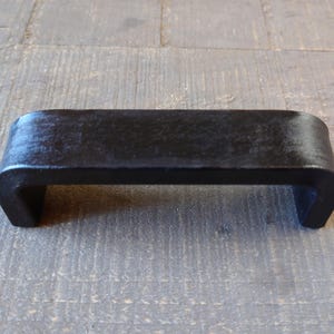 May include: A black, rectangular handle with curved sides and a flat top. The handle is made of a dark, possibly metal material, and is designed to be mounted on a surface. The handle is shown against a wooden background.
