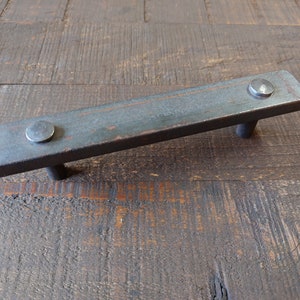 May include: A rustic, rectangular metal drawer pull with a dark gray finish and two silver rivets. The pull is shown on a wooden surface.
