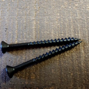 May include: Two black screws with a flat head and a Phillips drive. The screws are lying on a brown wooden surface.