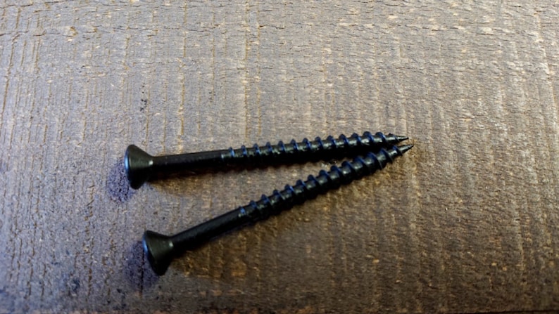 May include: Two black screws with a flat head and a Phillips drive. The screws are lying on a brown wooden surface.