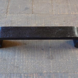 May include: A black, rectangular cabinet pull with a curved handle. The hardware is made of a dark, textured metal and is mounted on a wooden surface. The pull is approximately 8 inches long.