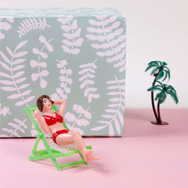 May include: A plastic figurine of a woman wearing a red bikini, relaxing on a green deckchair. The figurine is in front of a light green box with a white leaf pattern.