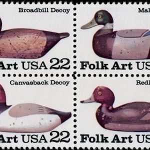 May include: Four vintage-style wooden duck decoys, each with a different species: Broadbill, Mallard, Canvasback, and Redhead. The decoys are painted in realistic colors and have a worn, rustic look. Each stamp features the text "Folk Art USA 22".