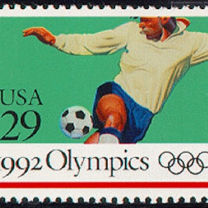 Puede incluir: Un sello postal conmemorativo de los Juegos Ol&iacute;mpicos de 1992 de color verde y blanco que representa a un jugador de f&uacute;tbol pateando un bal&oacute;n. El sello tiene el texto "USA 29" y los anillos ol&iacute;mpicos.