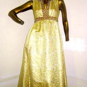 Puede incluir: Un vestido dorado sin mangas sobre un maniqu&iacute;. El vestido presenta un escote en V y un cintur&oacute;n con cuentas. La tela tiene un patr&oacute;n texturizado. El maniqu&iacute; tambi&eacute;n es dorado, con un peinado estilizado y labios rojos. El vestido es largo.
