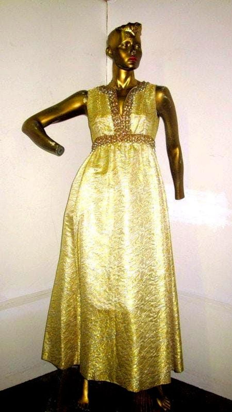 Puede incluir: Un vestido dorado sin mangas sobre un maniqu&iacute;. El vestido presenta un escote en V y un cintur&oacute;n con cuentas. La tela tiene un patr&oacute;n texturizado. El maniqu&iacute; tambi&eacute;n es dorado, con un peinado estilizado y labios rojos. El vestido es largo.