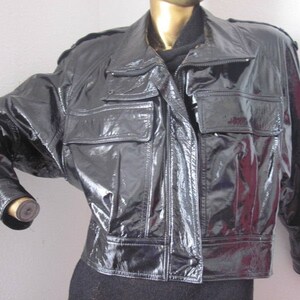 Pode incluir: Casaco preto brilhante com aspecto de couro envernizado. Possui gola, vários bolsos e fecho de correr. As mangas têm punhos abotoados e o casaco parece ser de estilo curto.