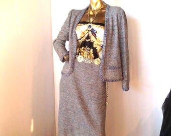 May include: A gold mannequin displays a gray tweed skirt suit with a matching jacket and a patterned top. The jacket has a blue trim. A gold chain belt and necklace complete the ensemble. The outfit is a classic, elegant style.
