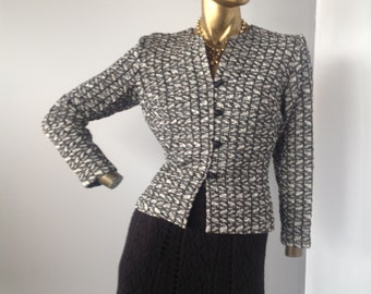 May include: A vintage two-piece outfit displayed on a mannequin. The jacket features a black and white geometric pattern and black buttons. The skirt is a long, black, open-weave knit. A gold necklace is visible.