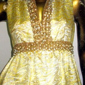 Puede incluir: Vestido amarillo sin mangas con un estampado arremolinado. El vestido presenta un escote en V y una cintura adornada con cuentas y pedrer&iacute;a doradas. El vestido se exhibe en un maniqu&iacute;.