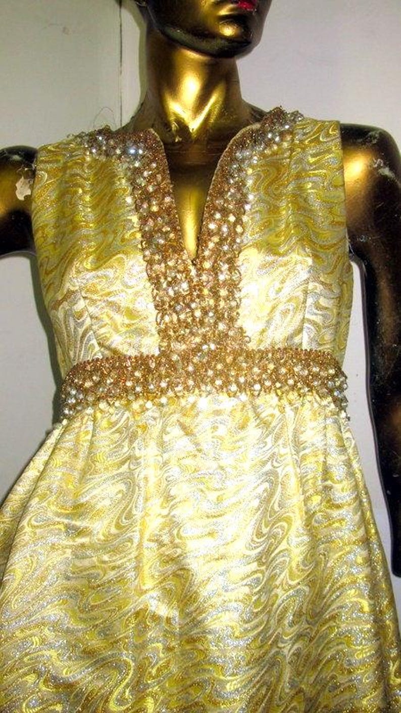 Puede incluir: Vestido amarillo sin mangas con un estampado arremolinado. El vestido presenta un escote en V y una cintura adornada con cuentas y pedrer&iacute;a doradas. El vestido se exhibe en un maniqu&iacute;.