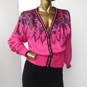 May include: A bright pink cardigan with a V-neck and long sleeves. The sweater features a black floral design on the shoulders and upper chest, with black trim along the neckline, cuffs, and button closure. The cardigan is buttoned up.