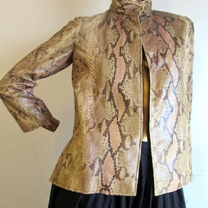 Puede incluir: Chaqueta con estampado de piel de serpiente y cuello alto. La chaqueta presenta una mezcla de tonos beige, marrón y rosa, imitando la piel de reptil. La chaqueta está abierta por delante y tiene mangas largas. La parte inferior de la imagen muestra una falda negra.