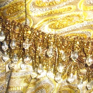Puede incluir: Primer plano de una tela brillante con un dise&ntilde;o de cachemira dorado y plateado. La tela est&aacute; adornada con un fleco de cuentas y gotas transparentes con forma de cristal, creando una apariencia lujosa y ornamentada. El tema general sugiere una prenda vintage o formal.