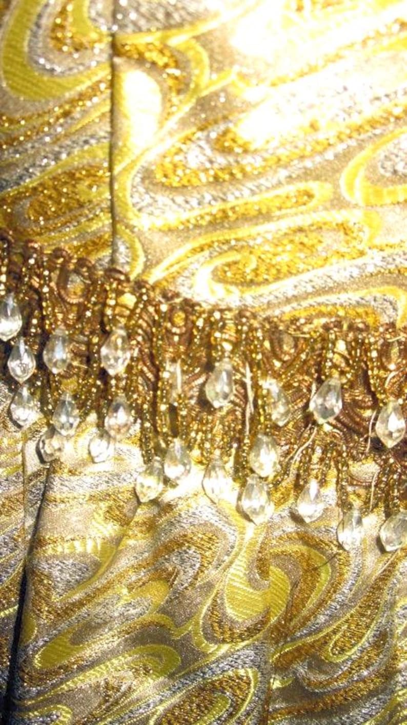 Puede incluir: Primer plano de una tela brillante con un dise&ntilde;o de cachemira dorado y plateado. La tela est&aacute; adornada con un fleco de cuentas y gotas transparentes con forma de cristal, creando una apariencia lujosa y ornamentada. El tema general sugiere una prenda vintage o formal.