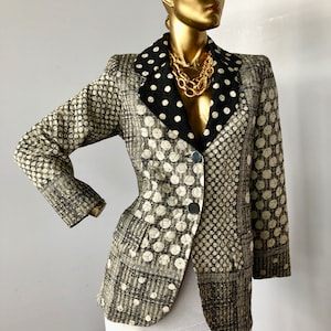 Puede incluir: Blazer estampado con cuello de lunares negros y collar de cadena dorada. La chaqueta presenta una mezcla de patrones geométricos y de puntos en tonos beige, negro y blanco. El blazer está abotonado y tiene un corte entallado.