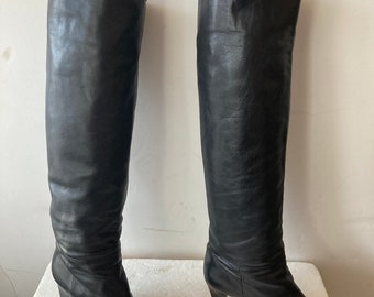 May include: A pair of black leather knee-high boots. The boots have a pointed toe and a small heel. The leather appears to be slightly creased. The boots are standing upright on a white surface against a white wall.
