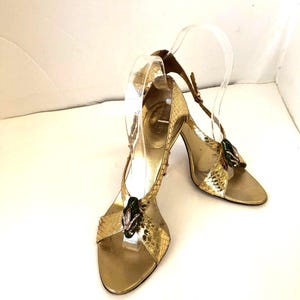 May include: A pair of gold high-heeled sandals featuring a textured snakeskin-like pattern. Each shoe has a T-strap design adorned with a dark green, oval-shaped decorative accent featuring small embedded crystals at the center and an ankle strap.