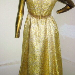 Puede incluir: Un vestido de fiesta dorado con un patr&oacute;n brillante y texturizado. El vestido presenta un escote y una cintura con cuentas, y una falda fluida. El vestido se exhibe en un maniqu&iacute; dorado.