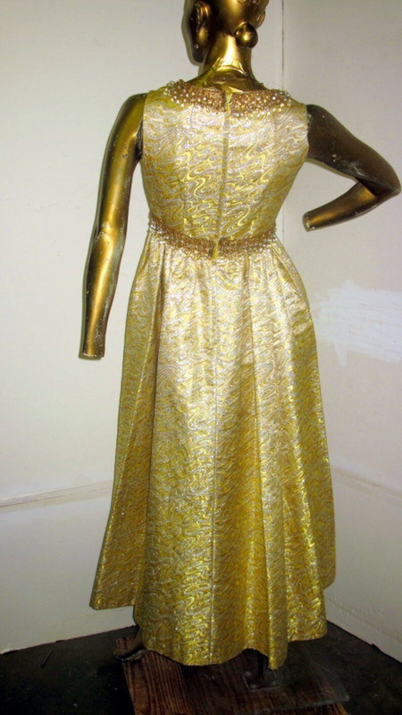 Puede incluir: Un vestido de fiesta dorado con un patr&oacute;n brillante y texturizado. El vestido presenta un escote y una cintura con cuentas, y una falda fluida. El vestido se exhibe en un maniqu&iacute; dorado.