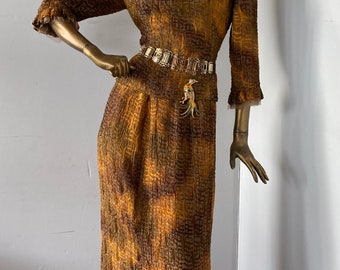 May include: A gold-painted mannequin wearing a long, textured brown and gold dress with a square neckline and long sleeves. The dress is accessorized with a decorative belt and brooches. The mannequin has a unique hairstyle and earrings.