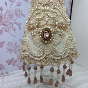 May include: A cream-colored fabric lampshade featuring intricate lace overlays, white pearl bead embellishments, and a central oval-shaped pink gemstone. The bottom edge is adorned with hanging clear crystal and metallic pink teardrop beads.