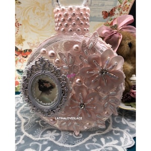 May include: A decorative, round glass bottle adorned with pink lace, pearls, and floral embellishments. A small framed cameo and the text "LATINALOVESLACE" are visible. The bottle's neck is embellished with pink pearls.