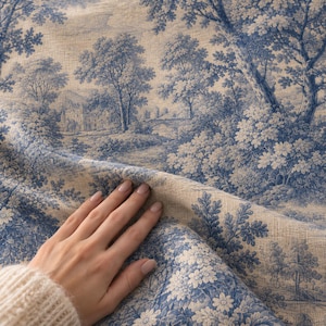 May include: A beige fabric featuring a blue toile de jouy pattern with detailed illustrations of trees, lush foliage, a stone bridge, and a large building in the background. The material has a visible woven texture and is draped in soft folds.