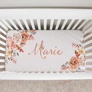 May include: A white crib with a fitted sheet featuring a floral design in shades of peach, brown, and gray. The sheet has the name "Marie" written in a cursive font in a copper color.