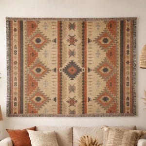 Puede incluir: Un tapiz tejido rectangular con un patrón geométrico en tonos beige, óxido, gris y negro. El tapiz tiene un borde con flecos y está colgado en una pared sobre un sofá con cojines a juego.