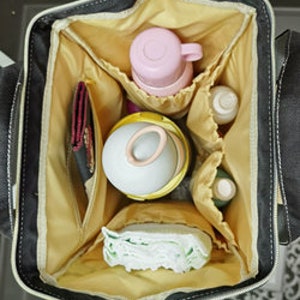 May include: A black diaper bag with a gold zipper and multiple pockets. The bag is open and filled with various baby items, including diapers, bottles, and a pink sippy cup.