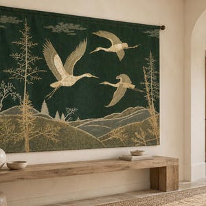 May include: A rectangular wall tapestry featuring a dark green background with a landscape scene. The design includes three flying cranes in light beige, stylized golden trees, and rolling hills. The tapestry is suspended from a dark metal rod.