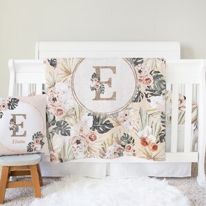 May include: A white crib with a floral patterned crib blanket and a matching pillow with the letter "E" and the name "Elaina" embroidered on it. The blanket and pillow feature a tropical theme with palm leaves, flowers, and a gold glitter letter "E" in a circle.