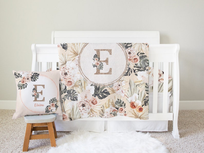 May include: A white crib with a floral patterned crib blanket and a matching pillow with the letter "E" and the name "Elaina" embroidered on it. The blanket and pillow feature a tropical theme with palm leaves, flowers, and a gold glitter letter "E" in a circle.