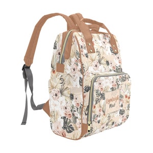 May include: A beige and brown floral print backpack with a brown leather trim and a zipper closure. The backpack has a large pocket on the front with the name "Lyndleigh Mae" printed on it.