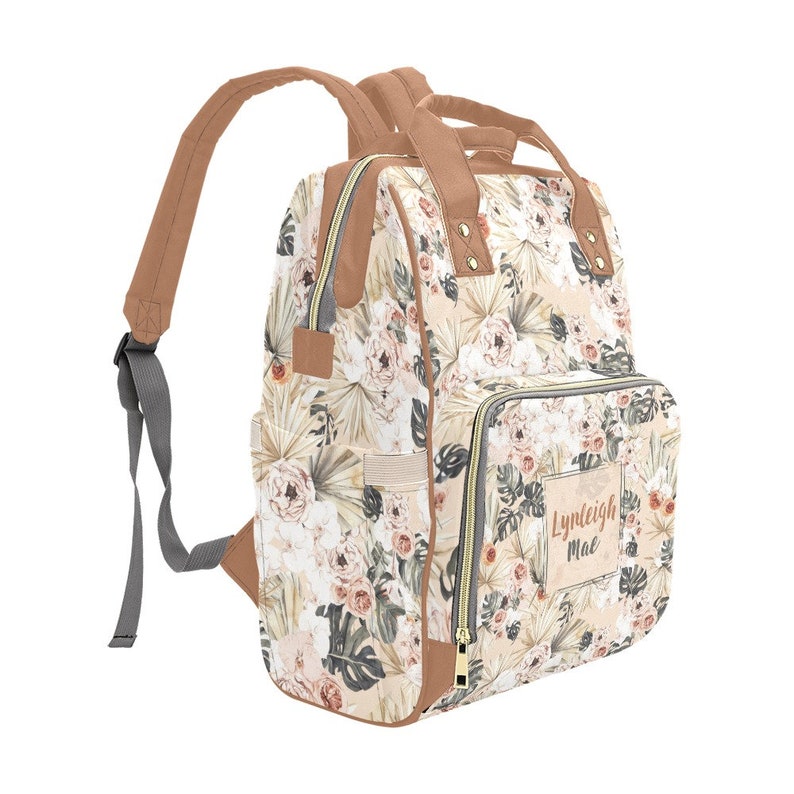 May include: A beige and brown floral print backpack with a brown leather trim and a zipper closure. The backpack has a large pocket on the front with the name "Lyndleigh Mae" printed on it.