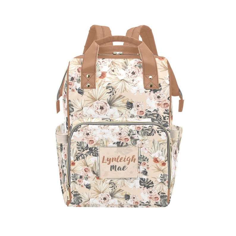 May include: A beige floral print diaper bag with brown straps and a zippered pocket. The bag has a personalized name tag that reads "Lynleigh Mae".