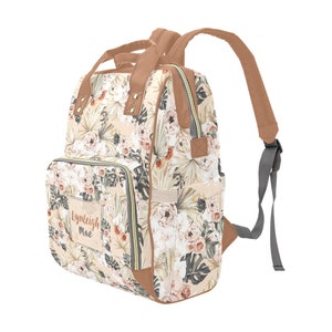 May include: A beige floral print diaper bag with brown faux leather accents and a personalized name tag that reads "Lynleigh Mae". The bag has a zippered front pocket, two side pockets, and a large main compartment. It also has adjustable straps for carrying on the back.