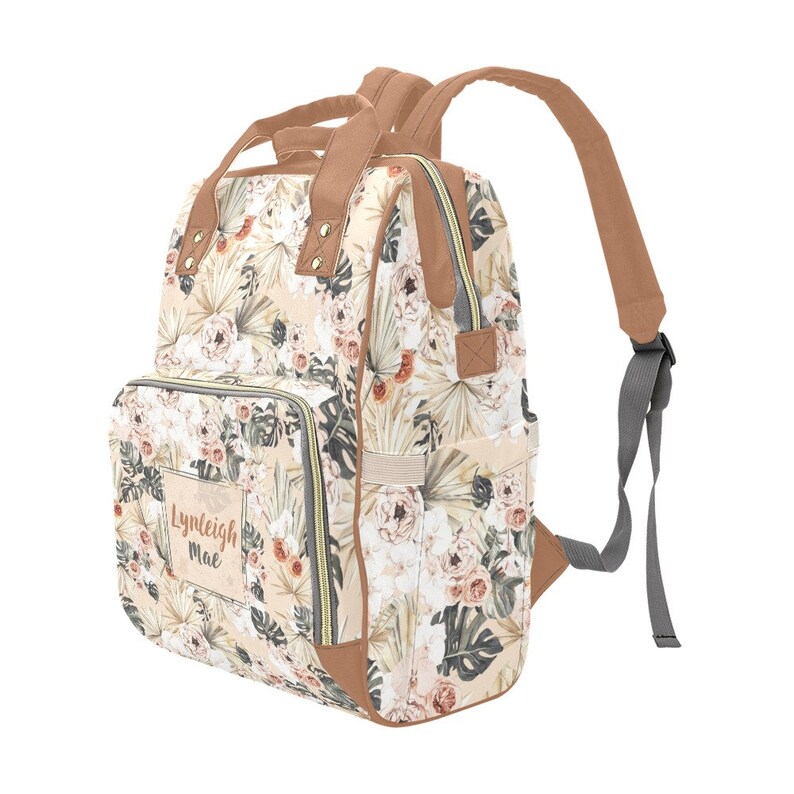 May include: A beige floral print diaper bag with brown faux leather accents and a personalized name tag that reads "Lynleigh Mae". The bag has a zippered front pocket, two side pockets, and a large main compartment. It also has adjustable straps for carrying on the back.