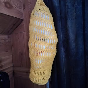 May include: A handmade, yellow crocheted storage bag, hanging from a metal bar. The bag has an open, lattice-like design, revealing white contents inside. The bag is suspended against a wooden wall and a dark blue curtain.