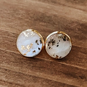 May include: A pair of round stud earrings with a gold-colored border. The earrings have a white background with gold leaf accents. The earrings are displayed on a wooden surface.